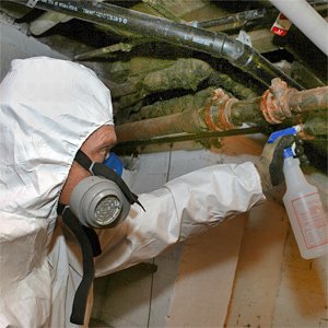 Worker in protective gear sprays a pipe with cleaning solution in a damp, enclosed space.