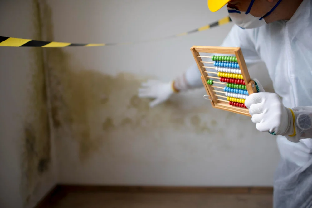 Person in protective gear measures mold on a wall with an abacus in a cordoned-off area.
