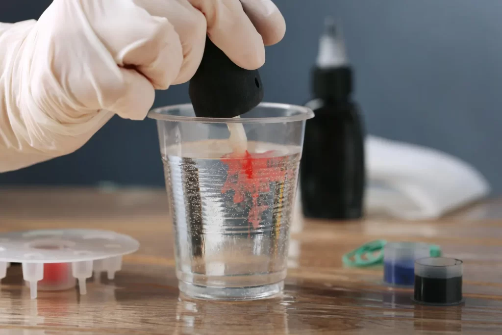 A gloved hand stirs a cup of water with red dye, surrounded by art supplies on a table.