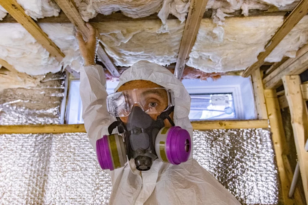 Worker in protective gear inspects insulation in a confined space with exposed beams.