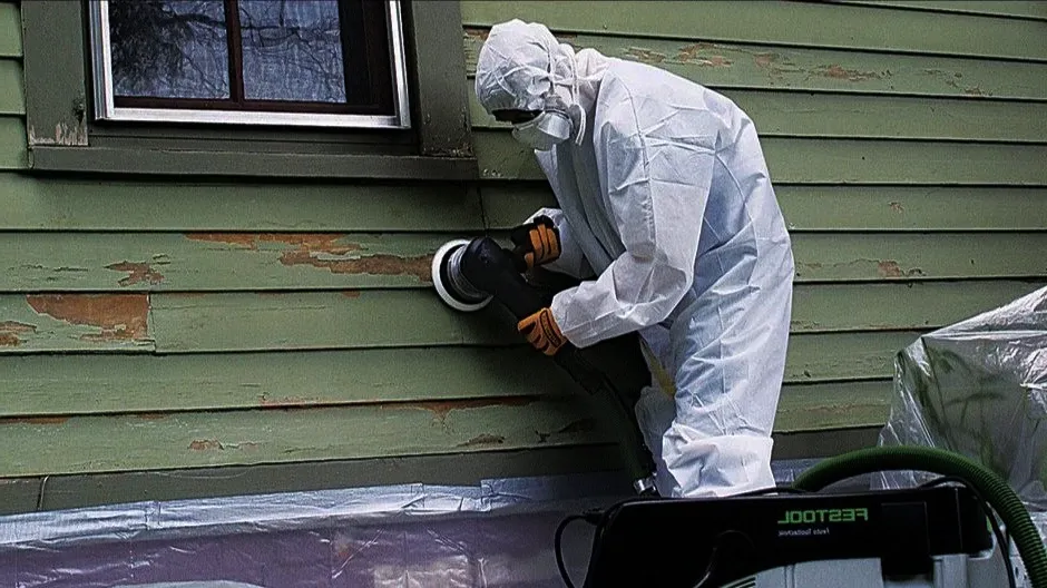 A worker in protective gear uses a sander on a peeling green wooden wall.