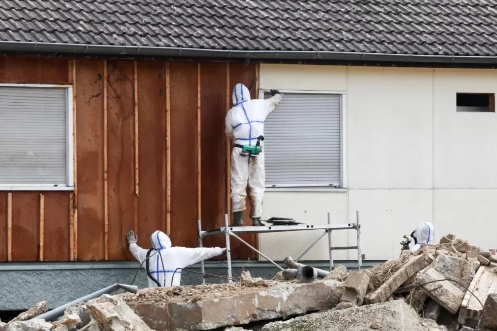 Workers in protective suits are renovating a building's exterior, with debris in the foreground.
