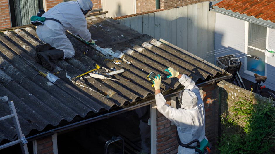Two workers in protective gear clean a roof, using tools to remove debris in bright sunlight.
